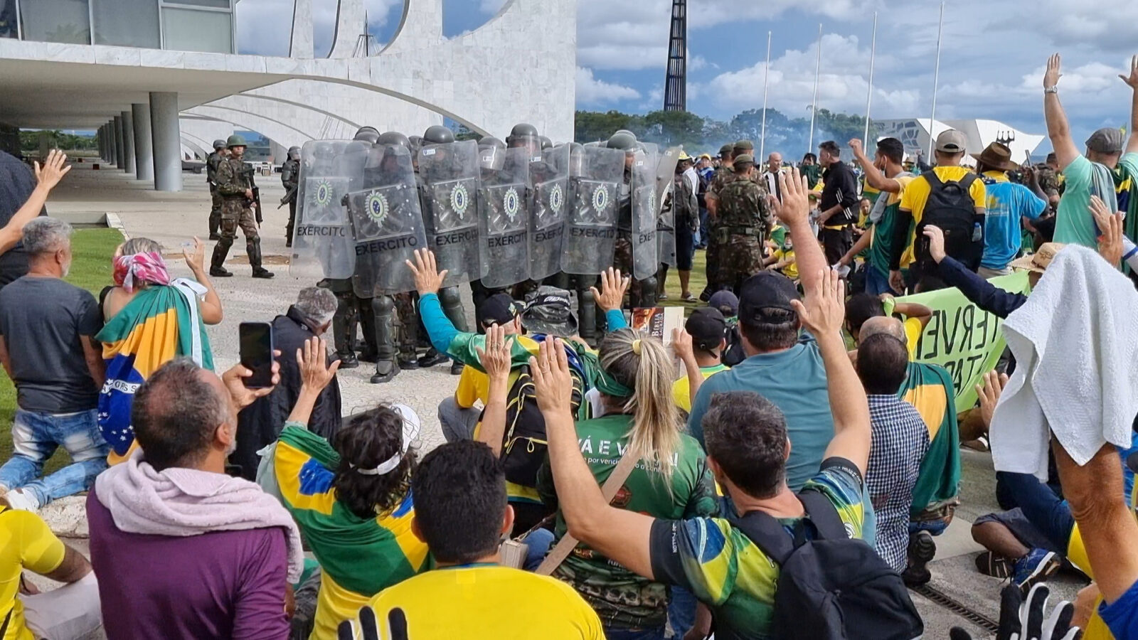 Bildet viser demonstranter med brasilianske flagg foran soldater med skjold merket "EXERCITO" utenfor en moderne bygning.