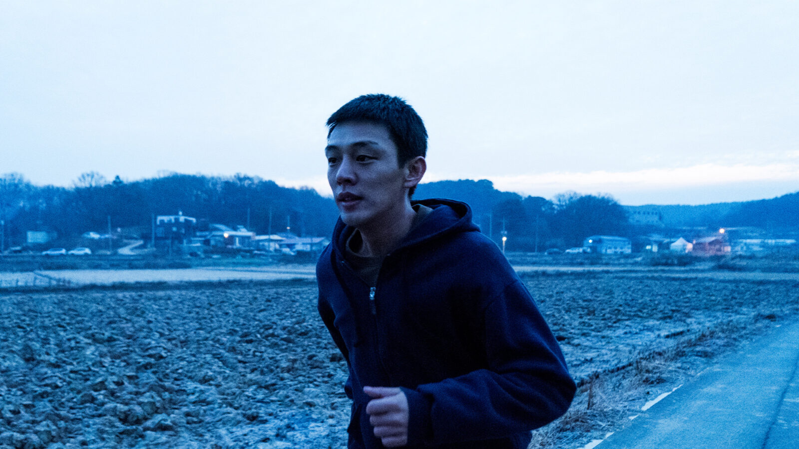 En ung mann jogger utendørs ved et jorde i skumringen, med bygninger og trær i bakgrunnen.