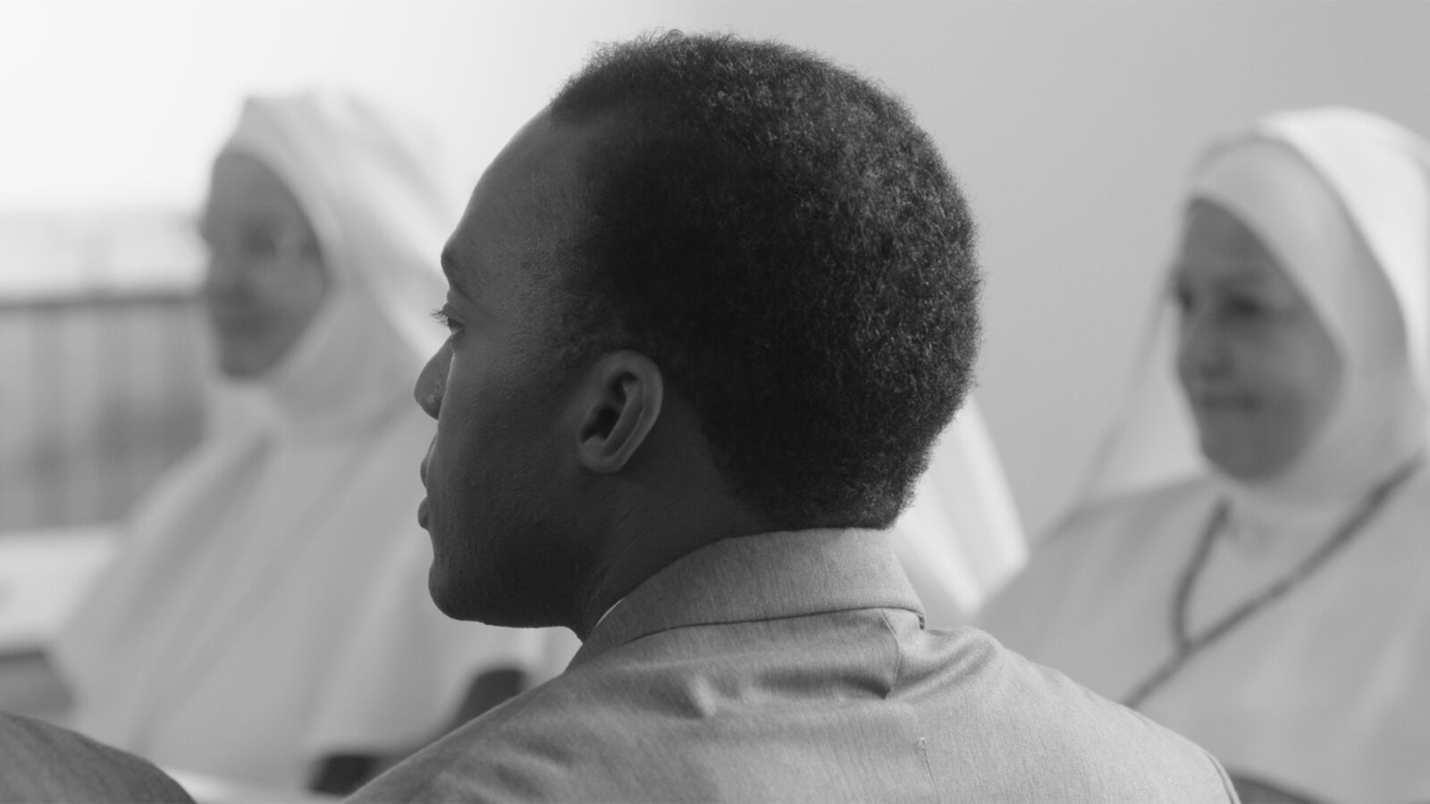 Frantz Fanon. Bildet viser en mann i dress, sett fra siden, med to nonner i hvite drakter i bakgrunnen.