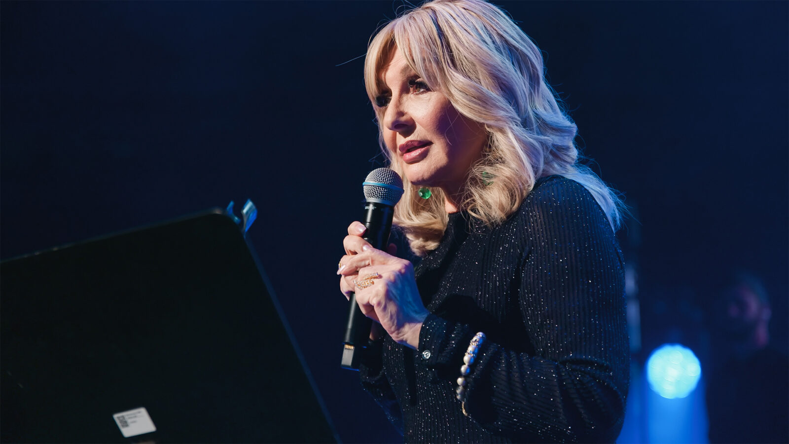 Googoosh. En kvinne med lyst hår, iført mørke klær og smykker, holder en mikrofon og står på en scene med mørk bakgrunn.