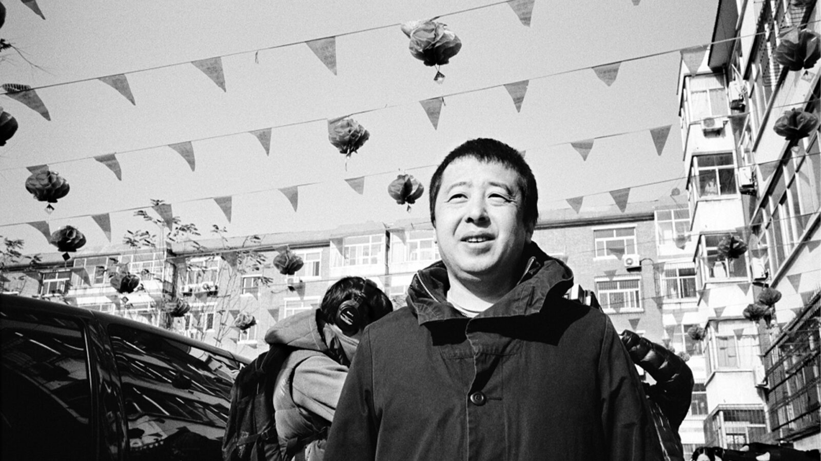 Jia Zhangke A Guy from Fenyang. Svart-hvitt bilde av en mann utendørs, omgitt av bygninger, flaggvimpler og dekorasjoner i luften.