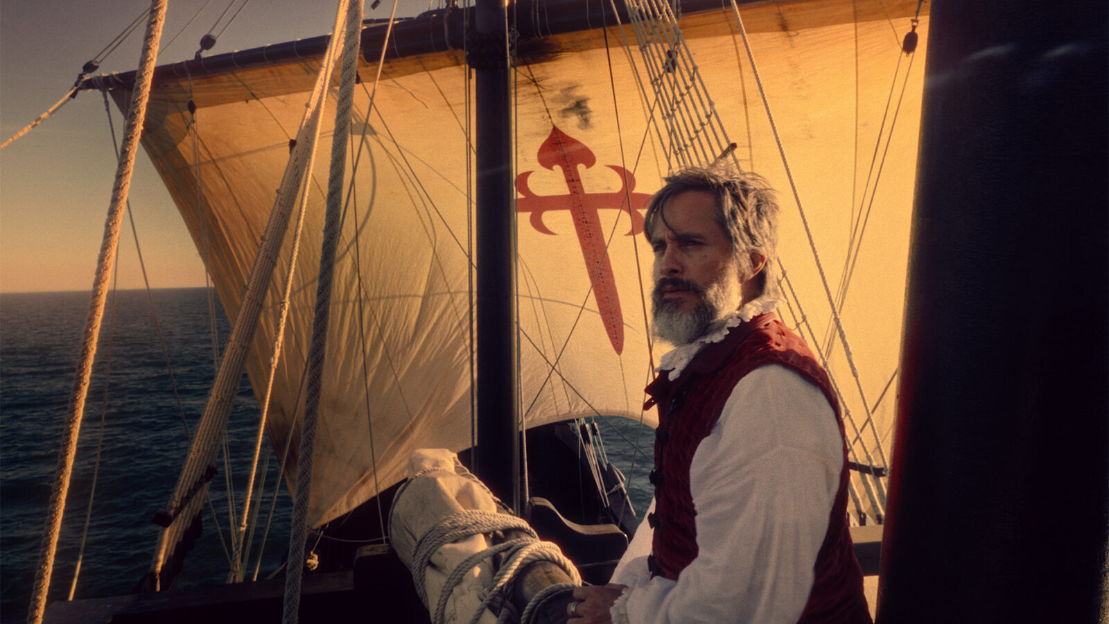 Magellan. En mann med skjegg og historiske klær står på et seilskip med et stort seil med rødt kors. Havet og solnedgang i bakgrunnen.