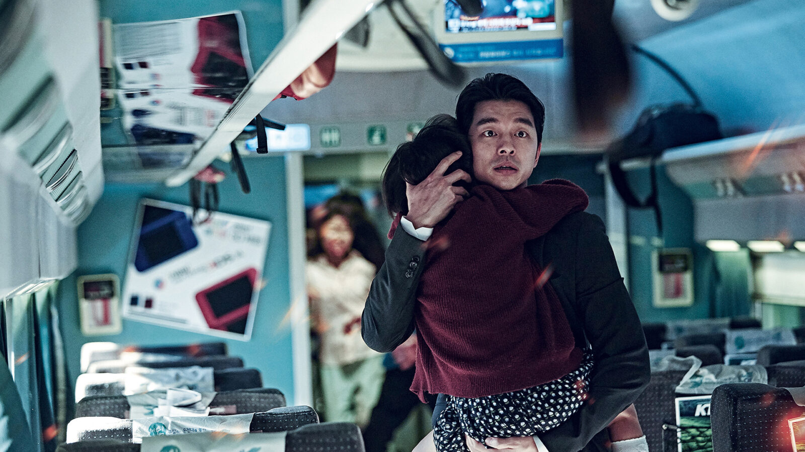 Train to Busan. En mann i dress holder et barn tett inntil seg inne i et tog. Flere personer og seter er synlige i bakgrunnen.