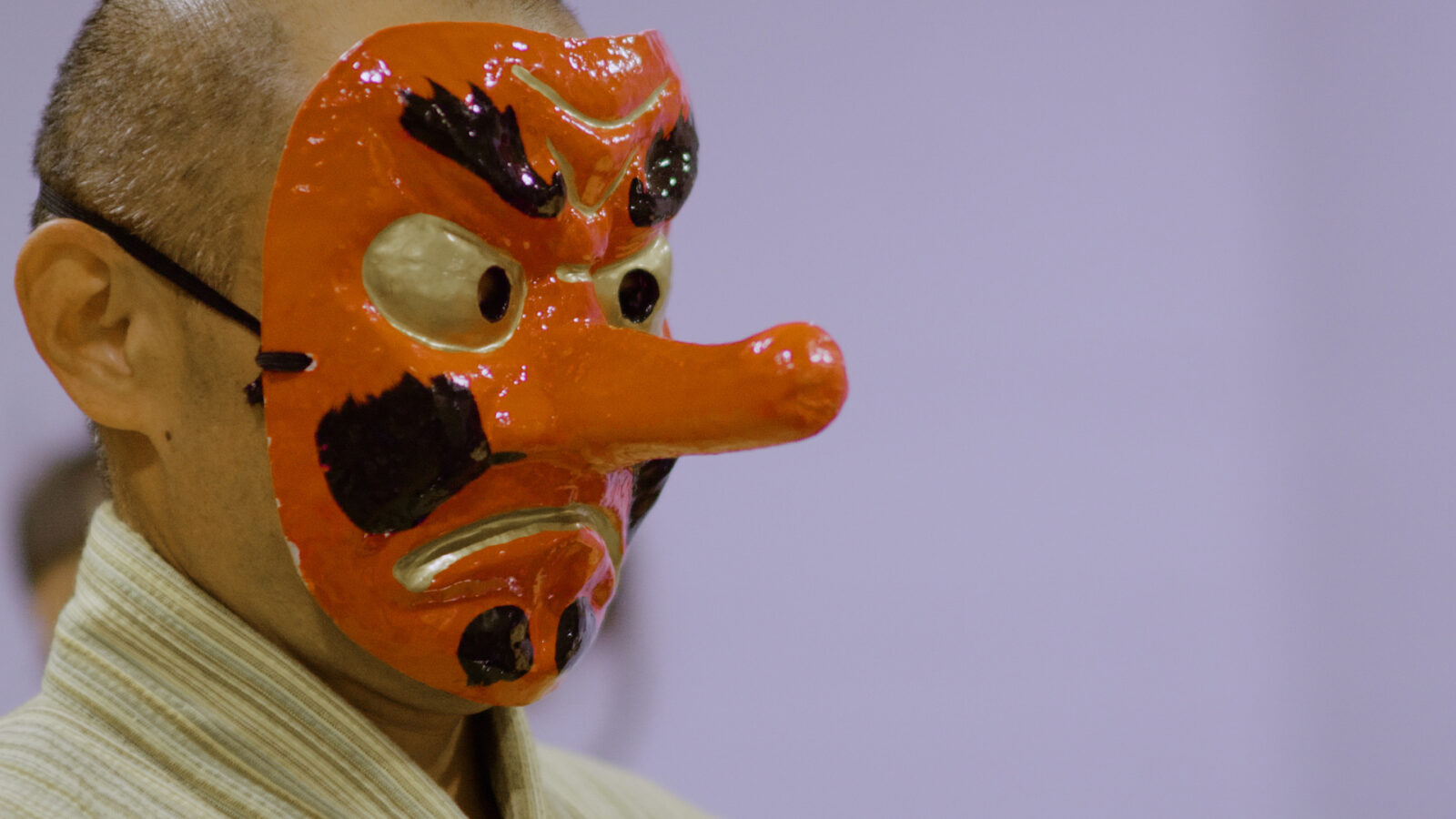 Bring Minyo Back En person med barbert hode har på seg en rød tradisjonell japansk maske med lang nese