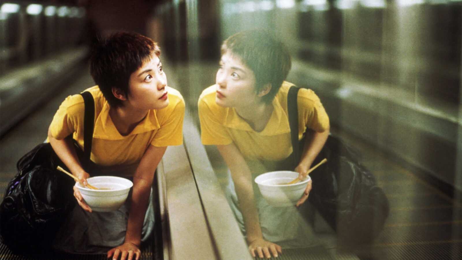 Chungking Express En kvinne med kort, moerkt haar og gul T-skjorte sitter paa huk i en rulletrapp med en bolle i haanden og ser oppover. Speilbildet hennes er synlig i glassveggen ved siden av henne.