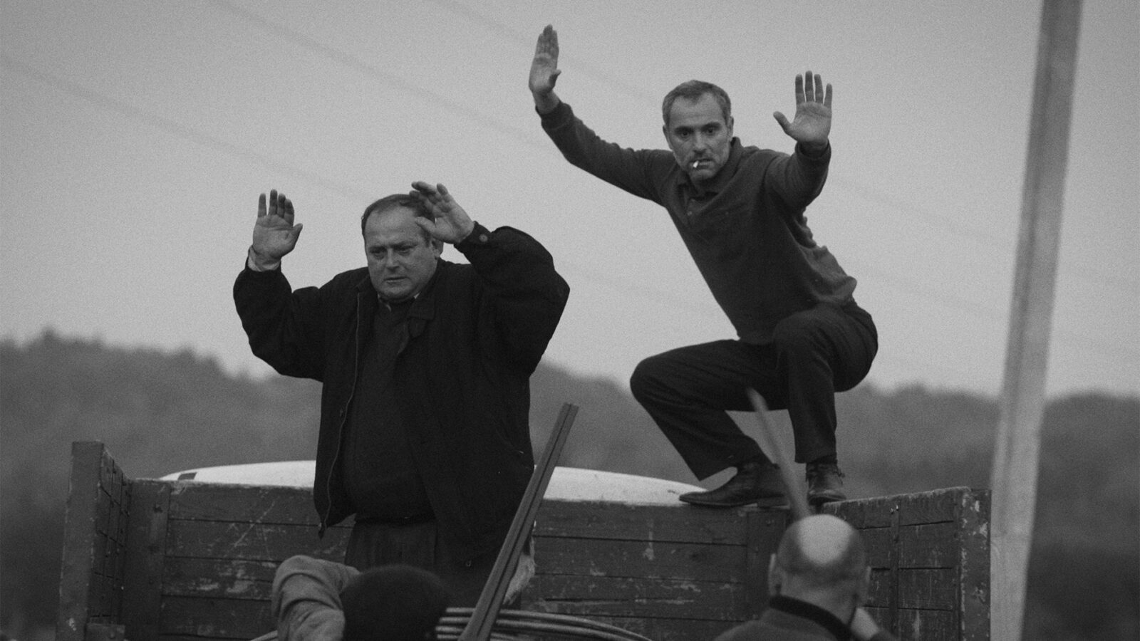 Guria To menn på et lasteplan med armene over hodet To personer peker paa dem med hagler Bildet er i svart hvitt