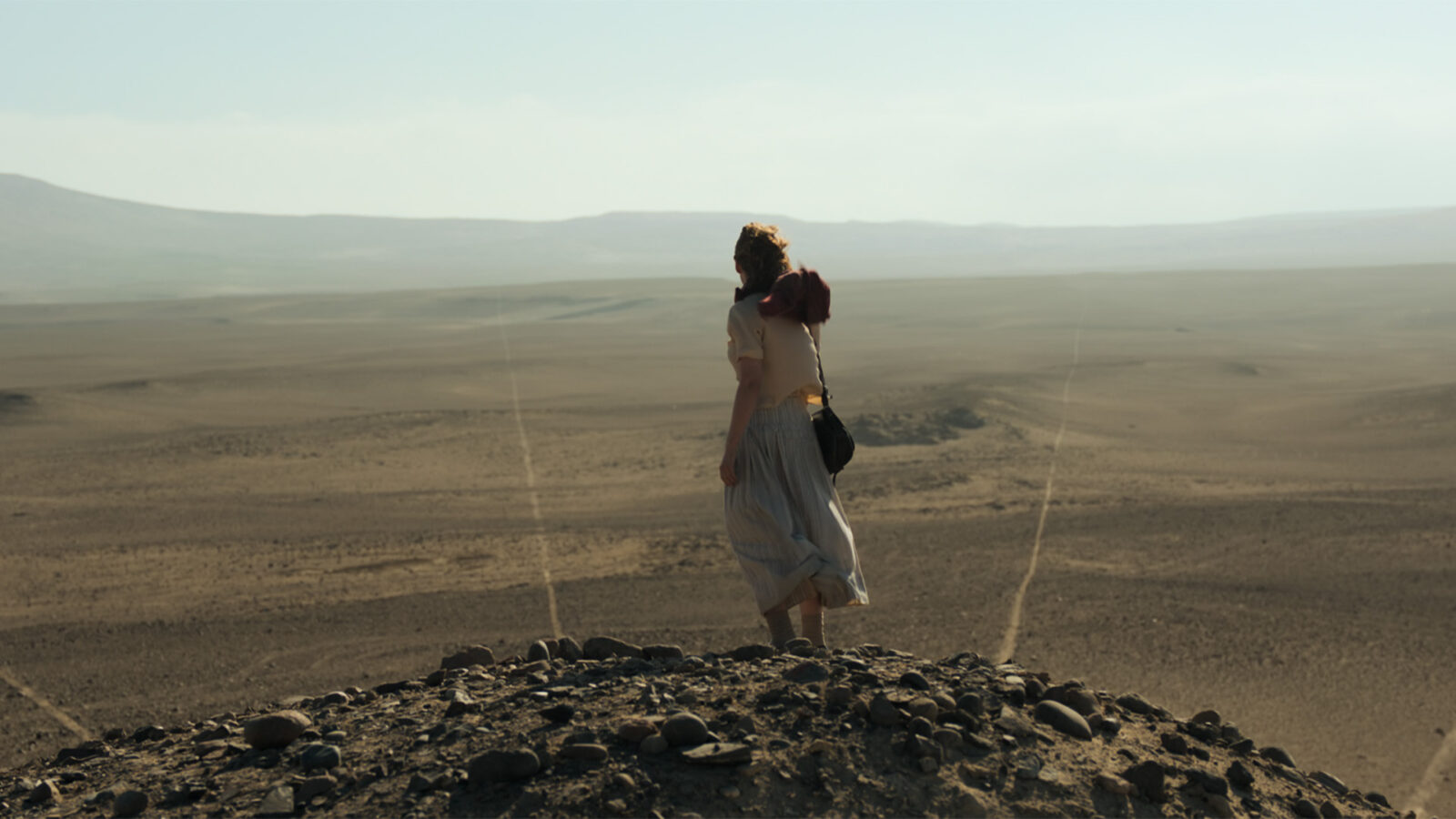 Lady Nazca En kvinne star pa en hoyde i en oyken med steiner foran seg og ser utover et flatt torket landskap med lange rette linjer i sanden