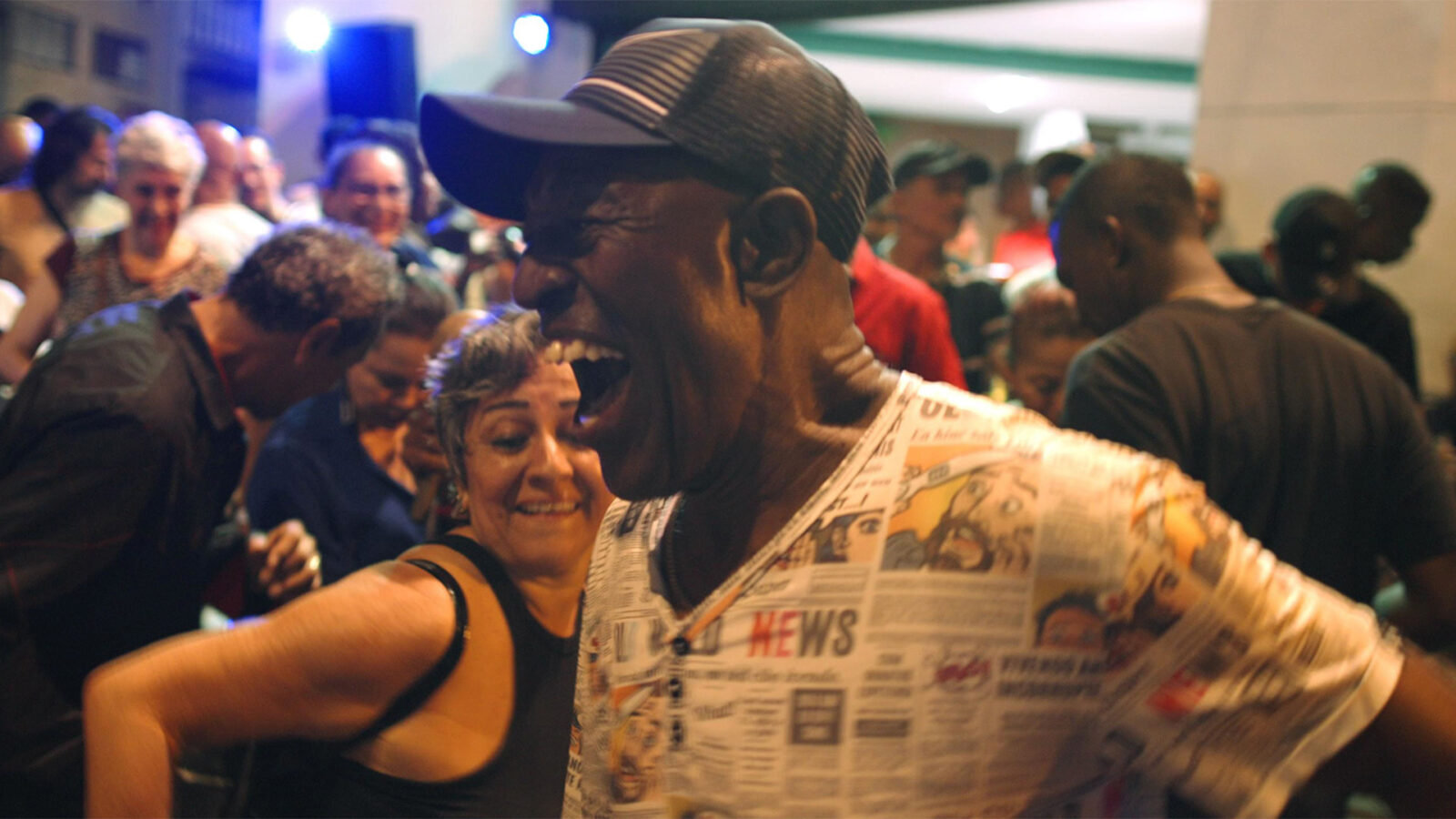 Salsa Lives En smilende mann med caps og T skjorte med avismotiv er i fokus Mange personer danser tett i bakgrunnen