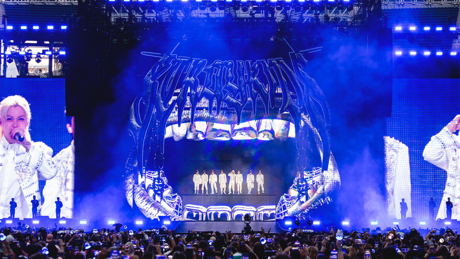 Stray Kids the Domin ATE Experience Stray Kids opptrer pa scene foran stort publikum Gruppen star foran stor skjerm med grafikk av tenner og bandnavn Alle har hvite antrekk