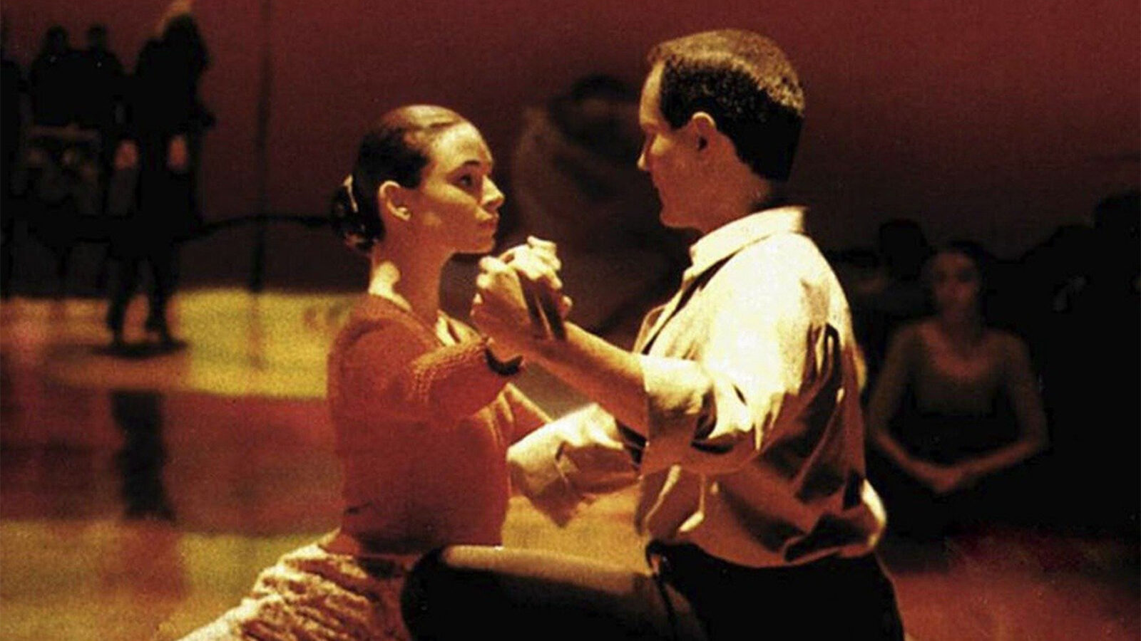 Tango To personer danser tett sammen i et rom med flere mennesker som ser på i bakgrunnen