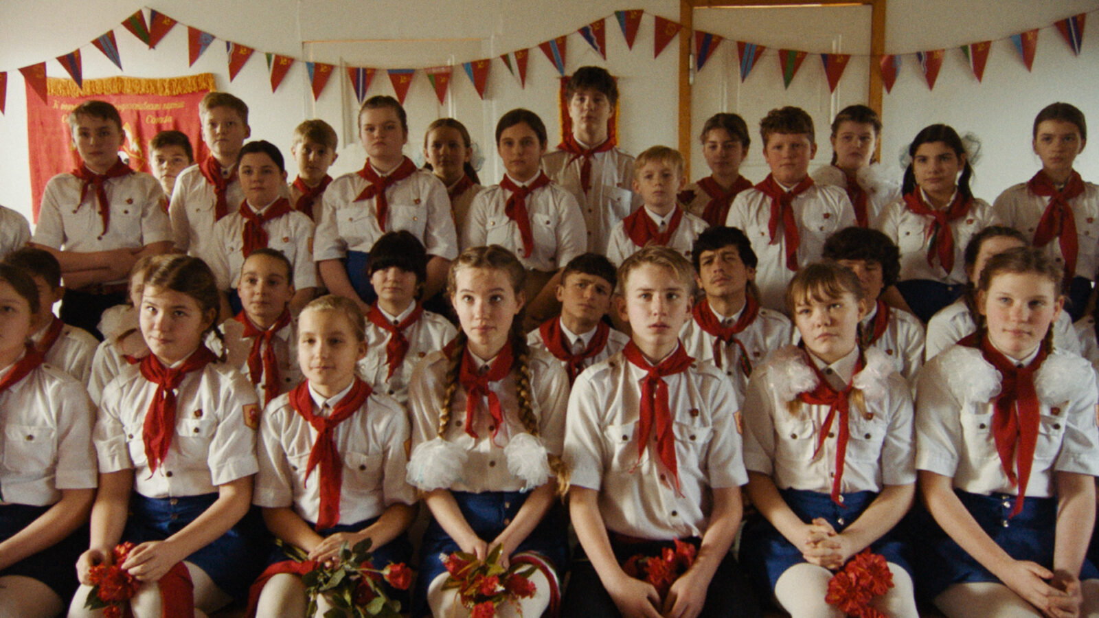 The Orphanage Bildet viser en gruppe barn i hvite skjorter bla skjort eller bukser og rode skjerf sittende i rekker foran dekorasjoner med sovjetiske symboler