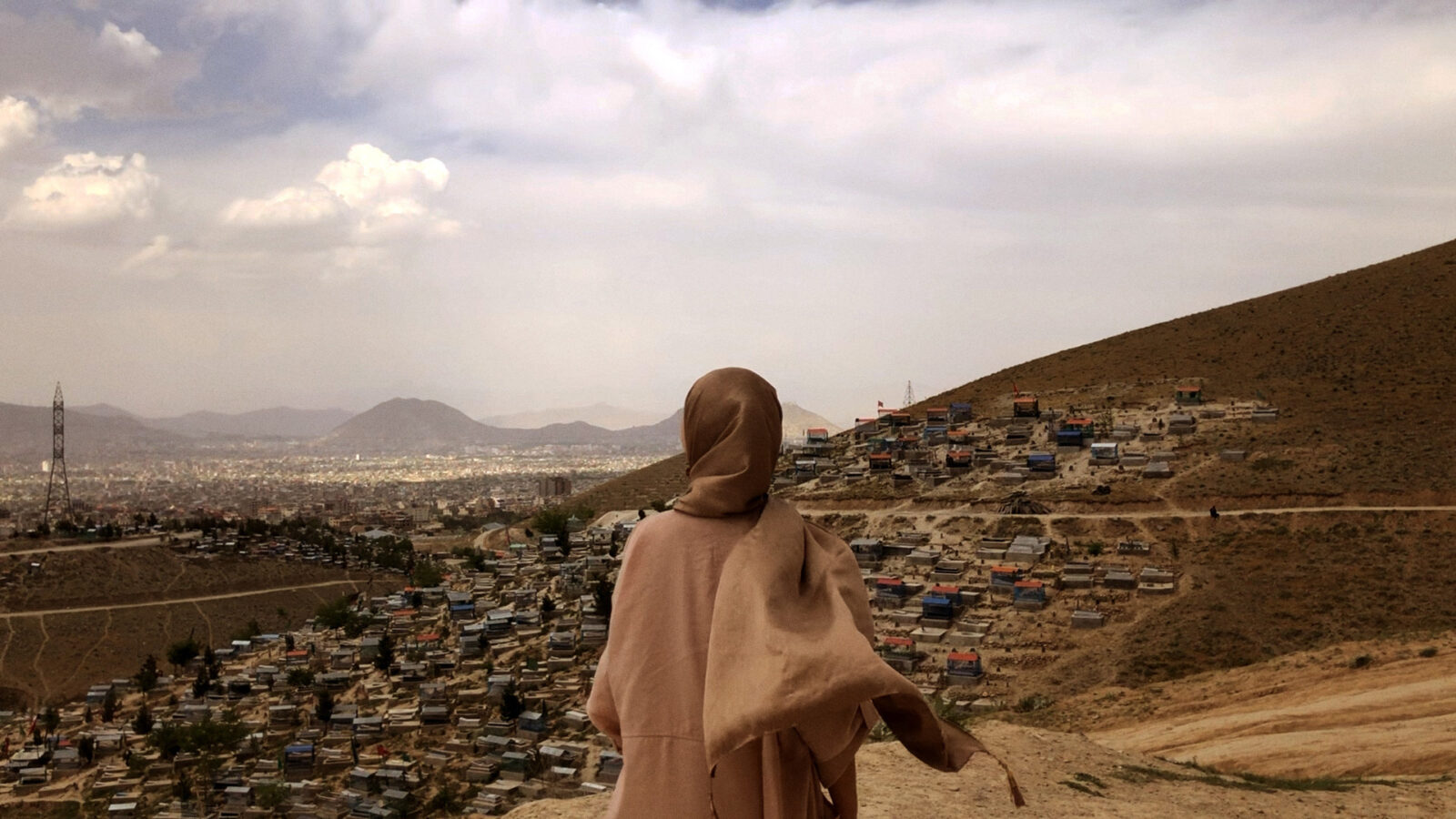 The Secret Reading Club of Kabul En person med sjal staar med ryggen til og ser utover en by og gravplasser i et omraade med bakker og fjell