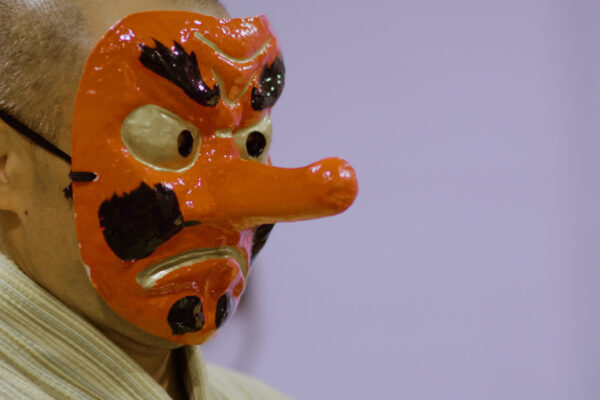 Bring Minyo Back En person med barbert hode har på seg en rød tradisjonell japansk maske med lang nese