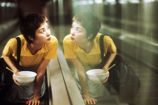 Chungking Express En kvinne med kort, moerkt haar og gul T-skjorte sitter paa huk i en rulletrapp med en bolle i haanden og ser oppover. Speilbildet hennes er synlig i glassveggen ved siden av henne.
