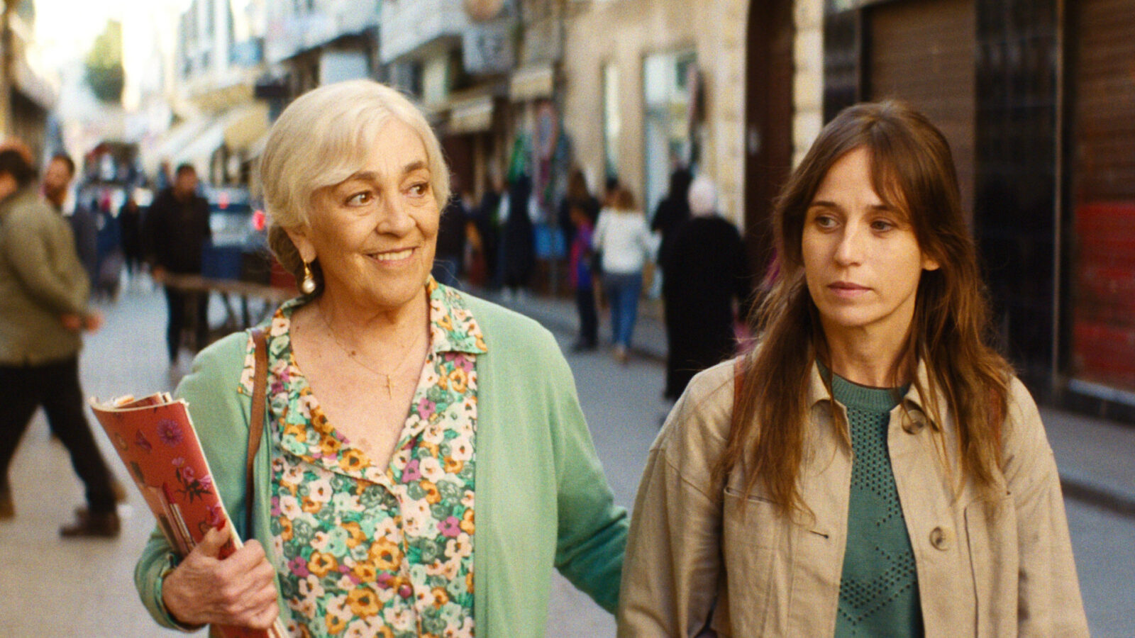 Calle Malaga To kvinner går pa en gate mellom bygninger Den ene har kort grått har og grønn jakke og holder en bok Den andre har langt brunt har og beige jakke