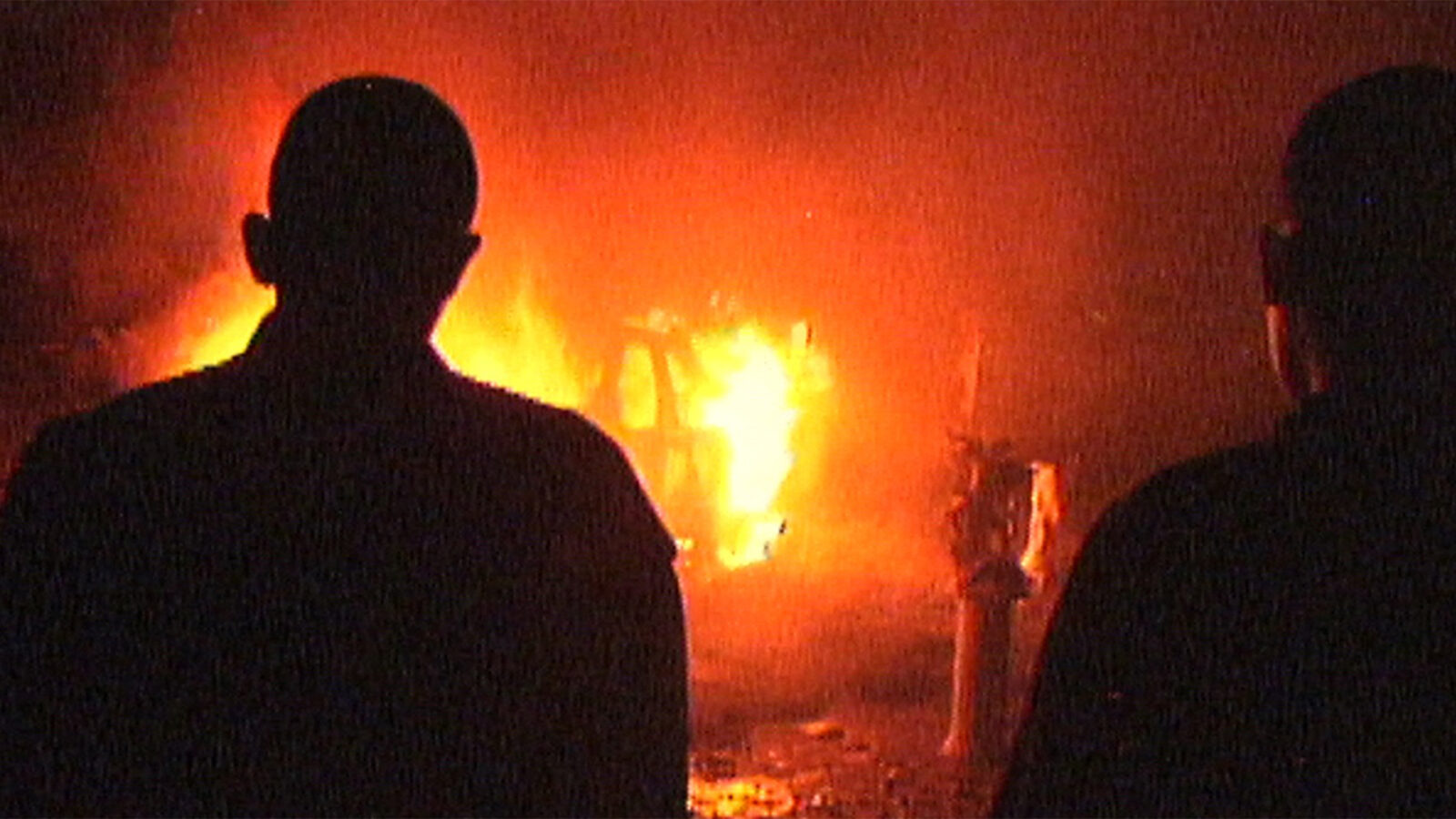 Barrio Triste To personer i silhuett står foran en stor flamme og brennende gjenstander Bakgrunnen er mørk Flammene lyser opp midten av bildet