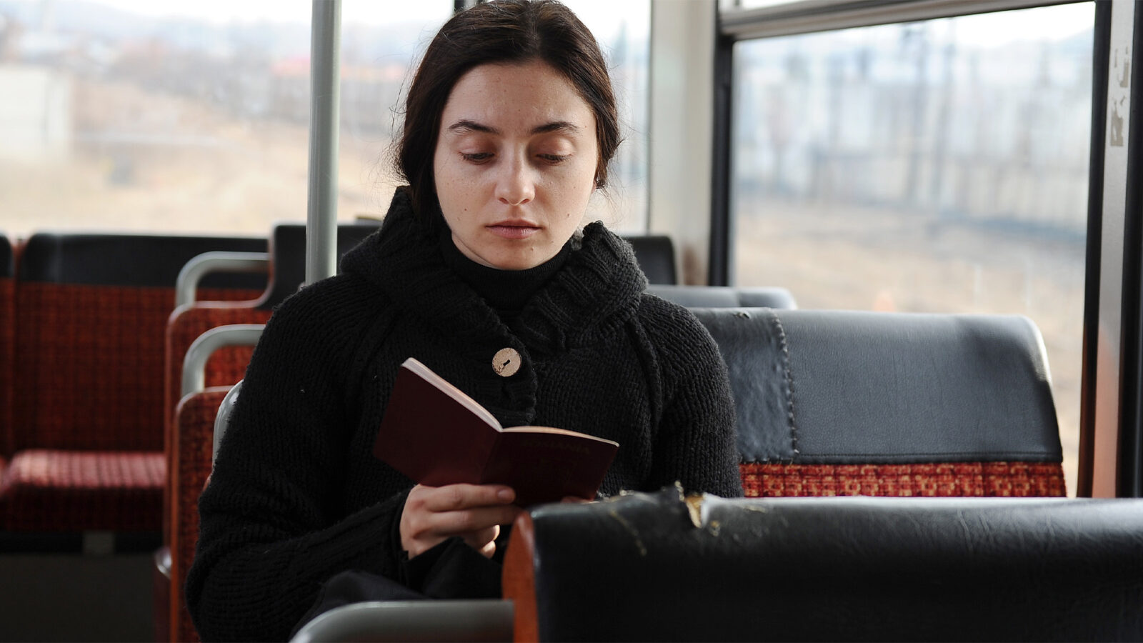 Beyond the Hills En kvinne med mørkt hår og svart genser sitter alene på et busssete og leser i en liten bok Bussens seter er svarte og røde