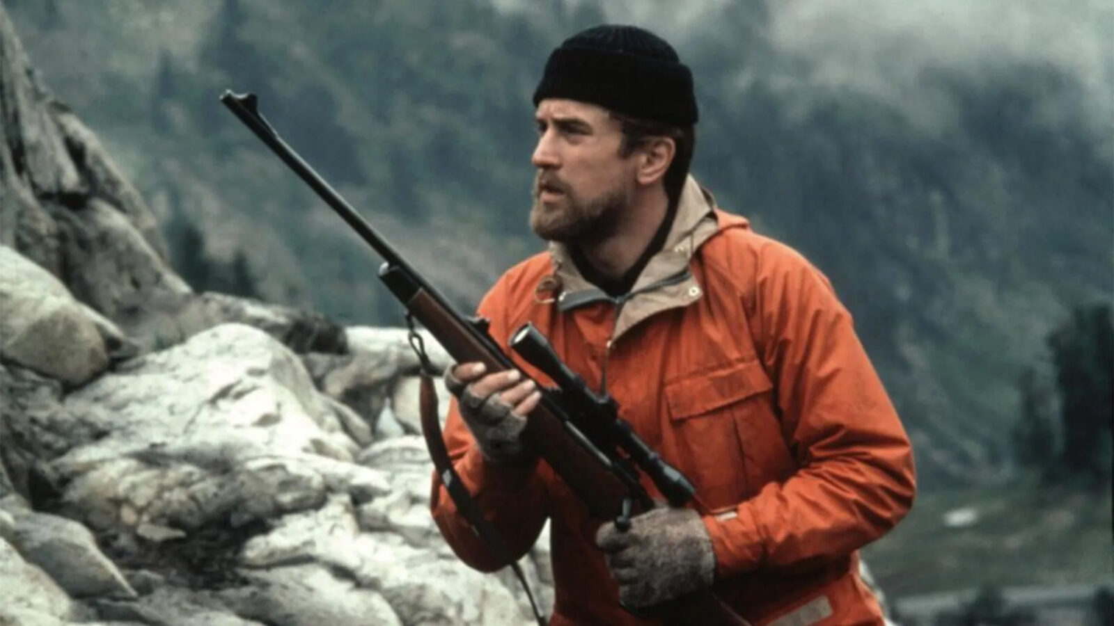 Hjortejegeren En mann med skjegg og oransje jakke holder et gevær Han har svart lue og grå votter Bak ham er det steiner og fjell