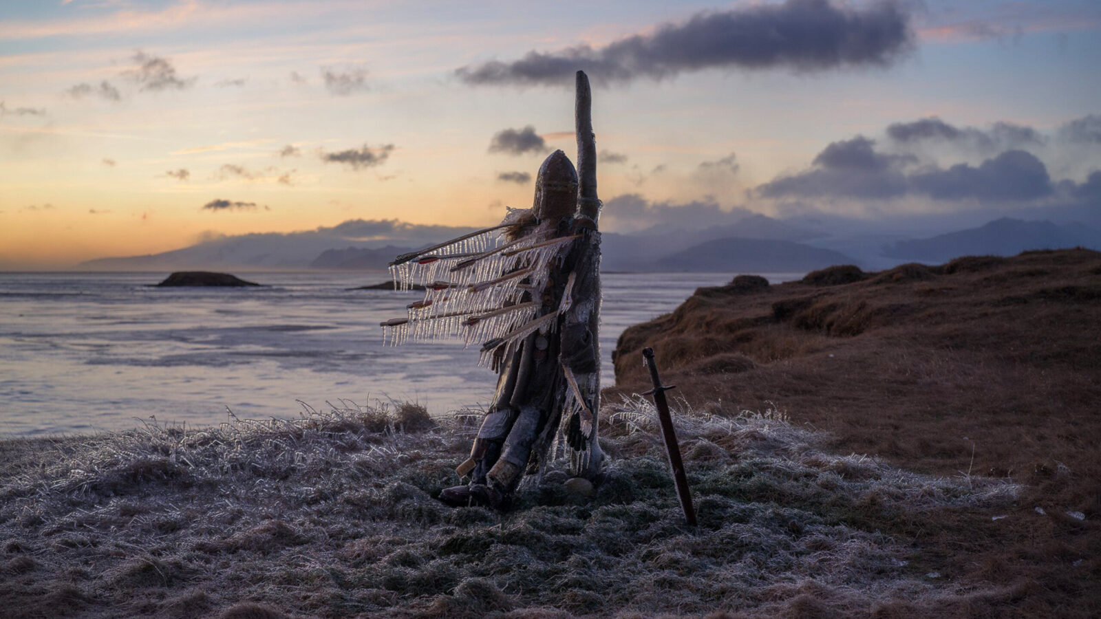 Joan of Arc En statue av en person i rustning med is på seg står på en gresslette ved vannet Et sverd står i bakken ved siden av Bakgrunnen viser fjell og himmel
