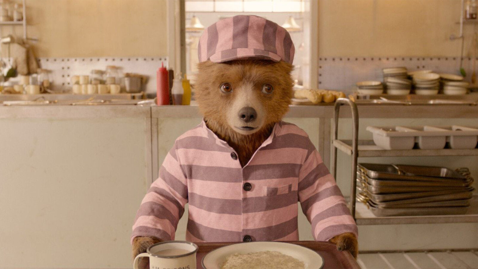 Paddington 2 En brun bjørn med stripete rosa og grå klær og lue står på et kjøkken og holder et brett med en tallerken og et krus