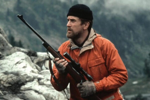 Hjortejegeren En mann med skjegg og oransje jakke holder et gevær Han har svart lue og grå votter Bak ham er det steiner og fjell