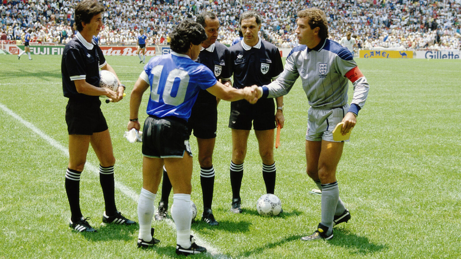To fotballspillere haandhilser foran dommere paa en gressbane med publikum i bakgrunnen