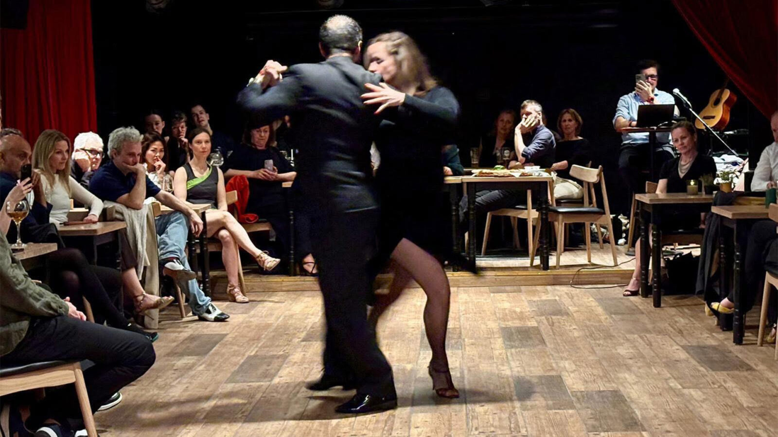 Tangokurs paa cinemateket. To personer danser tango paae tregulv foran publikum ved bord.