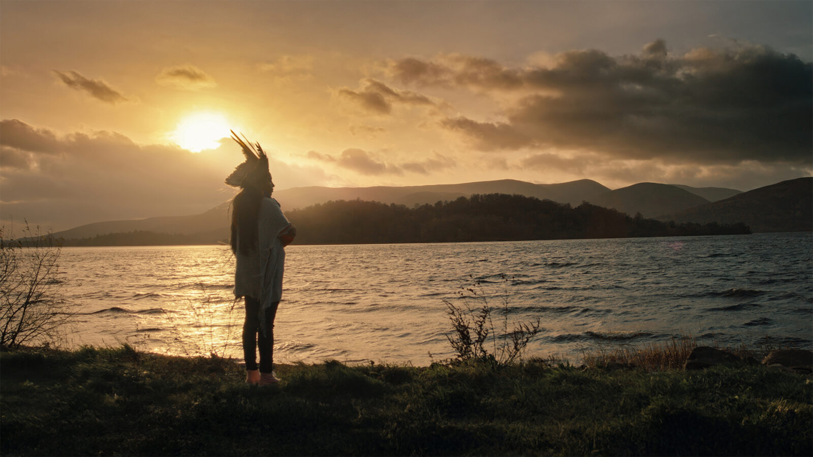 DKR Yanuni. En person med fjærpryd står ved en innsjø omgitt av fjell og natur i solnedgang.