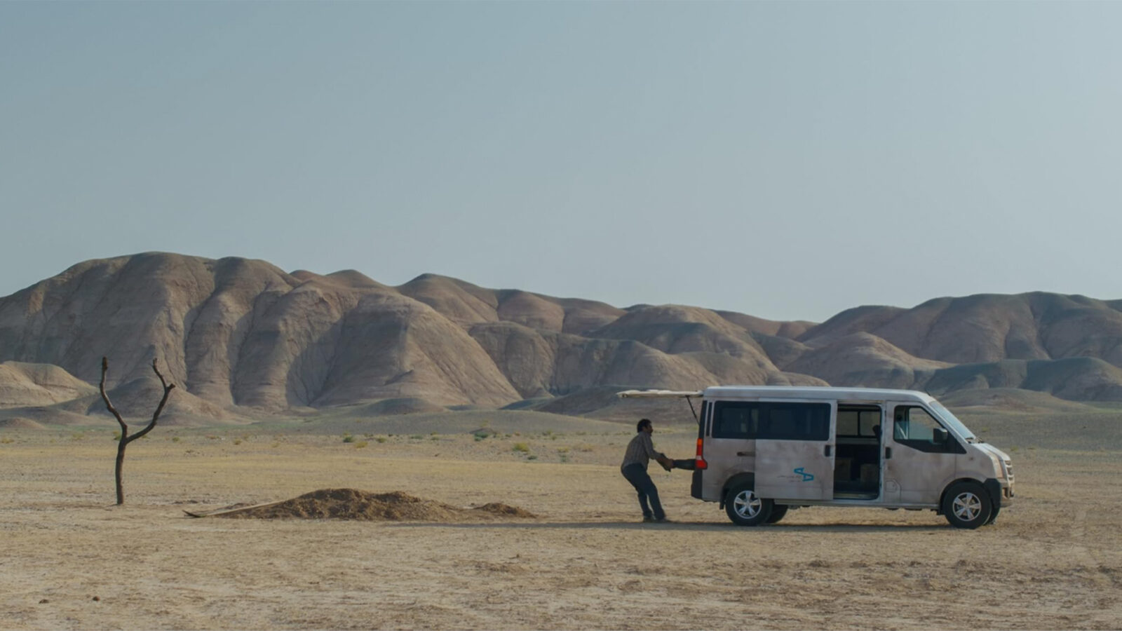 Jafar Panahi Regissørsamtale. En mann står bak en hvit varebil parkert i et tørt, ørkenlignende landskap. I bakgrunnen ligger avrundede sandfargede fjell, og et enslig, nakent tre står til venstre i bildet.