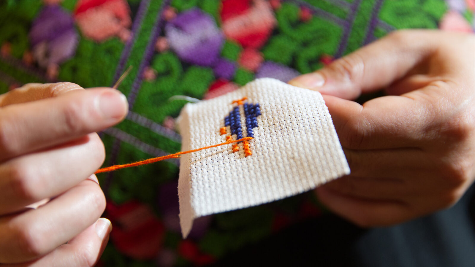 Kulturhub Tatreez og te Palestinsk broderiverksted Hender holder broderi med oransje og blått garn I bakgrunnen er det et grønt rødt og lilla mønster