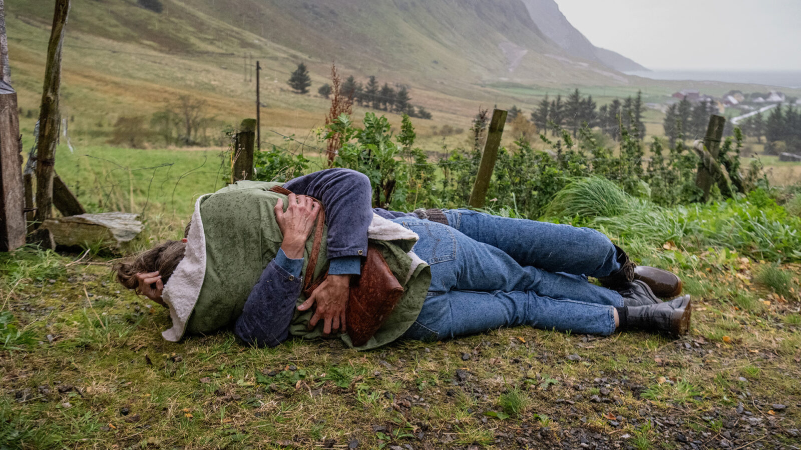 Kjærast på det norske teatret To personer ligger på bakken utendørs En person i grønn jakke og en i blå genser og jeans Fjell og grønt landskap i bakgrunnen